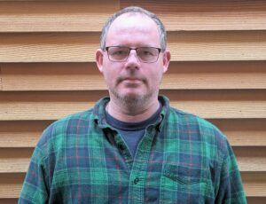 Man wearing glasses and a plaid shirt, standing in front of wood paneling.