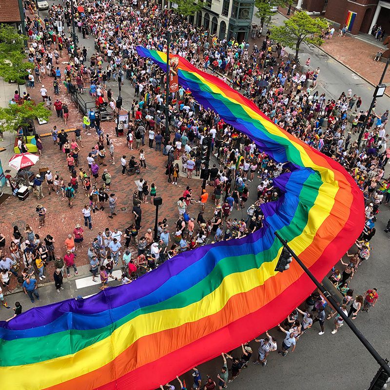 Large rainbow flag unfurls at a Pride parade, surrounded by a crowd of people in a city setting.