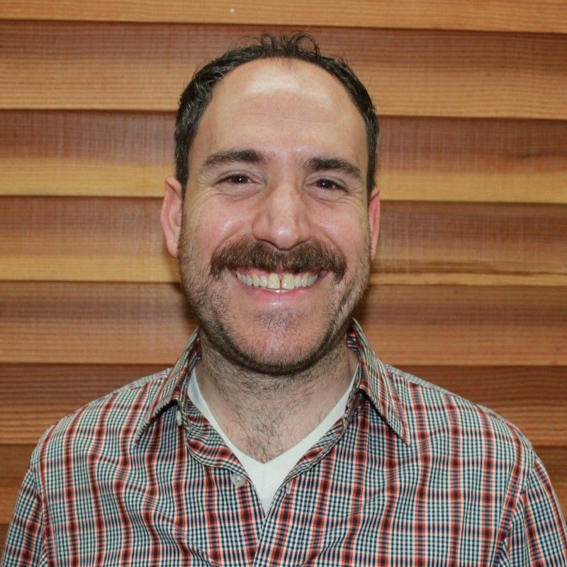 Man with brown hair and mustache smiles, wearing a plaid shirt against a wood-paneled wall.