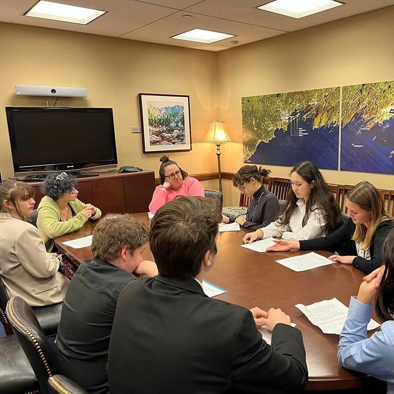 Group of people seated around a table, documents present, engaged in a discussion. Interior setting with artwork and a map on wall.