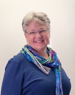 Woman with short grey hair and glasses smiles, wearing blue shirt and colorful scarf.