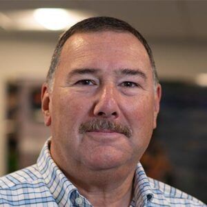 Man with a mustache wearing a plaid shirt, smiling in an office setting.