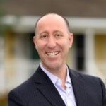 Man with a bald head in a suit smiling broadly, with a light blue shirt. Blurred background of a building.