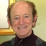Smiling man with reddish-brown hair in a maroon sweater, indoors in front of a painting.