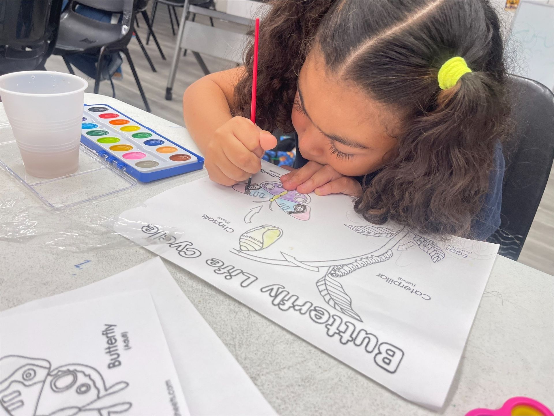 A little girl is sitting at a table painting a picture of a butterfly.