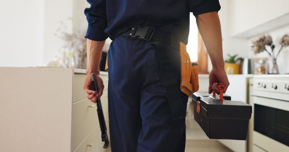 Person in navy work clothes walks toward a kitchen, carrying a toolbox and tool.