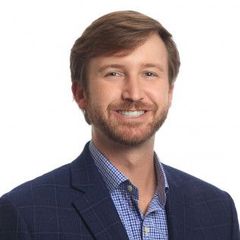 Man with brown hair and beard, wearing a blue blazer over a checkered button-down, smiling.