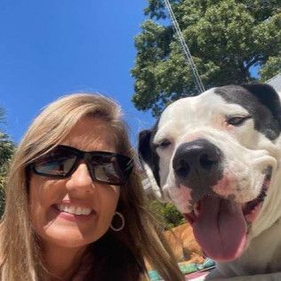 Woman with sunglasses smiles beside a black and white dog with tongue out, sunny outdoors.