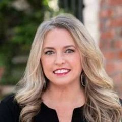 Woman with blonde hair smiling, wearing black top, posing outdoors near brick building.