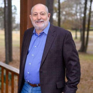 Man in a blue shirt, jeans, and a checkered blazer smiles, leaning on a porch railing with trees in the background.