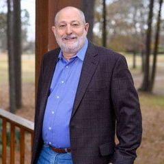 Man in a blue shirt, jeans, and a checkered blazer smiles, leaning on a porch railing with trees in the background.