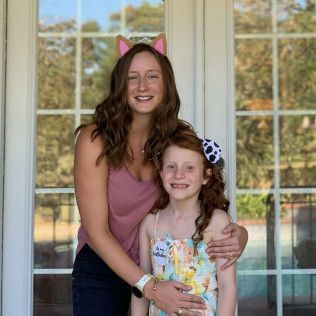 Woman and child smiling, embracing outdoors. The woman wears pink cat ears, the child has a cow-print bow.