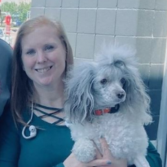 Woman smiling, holding a fluffy gray and white poodle with orange collar.