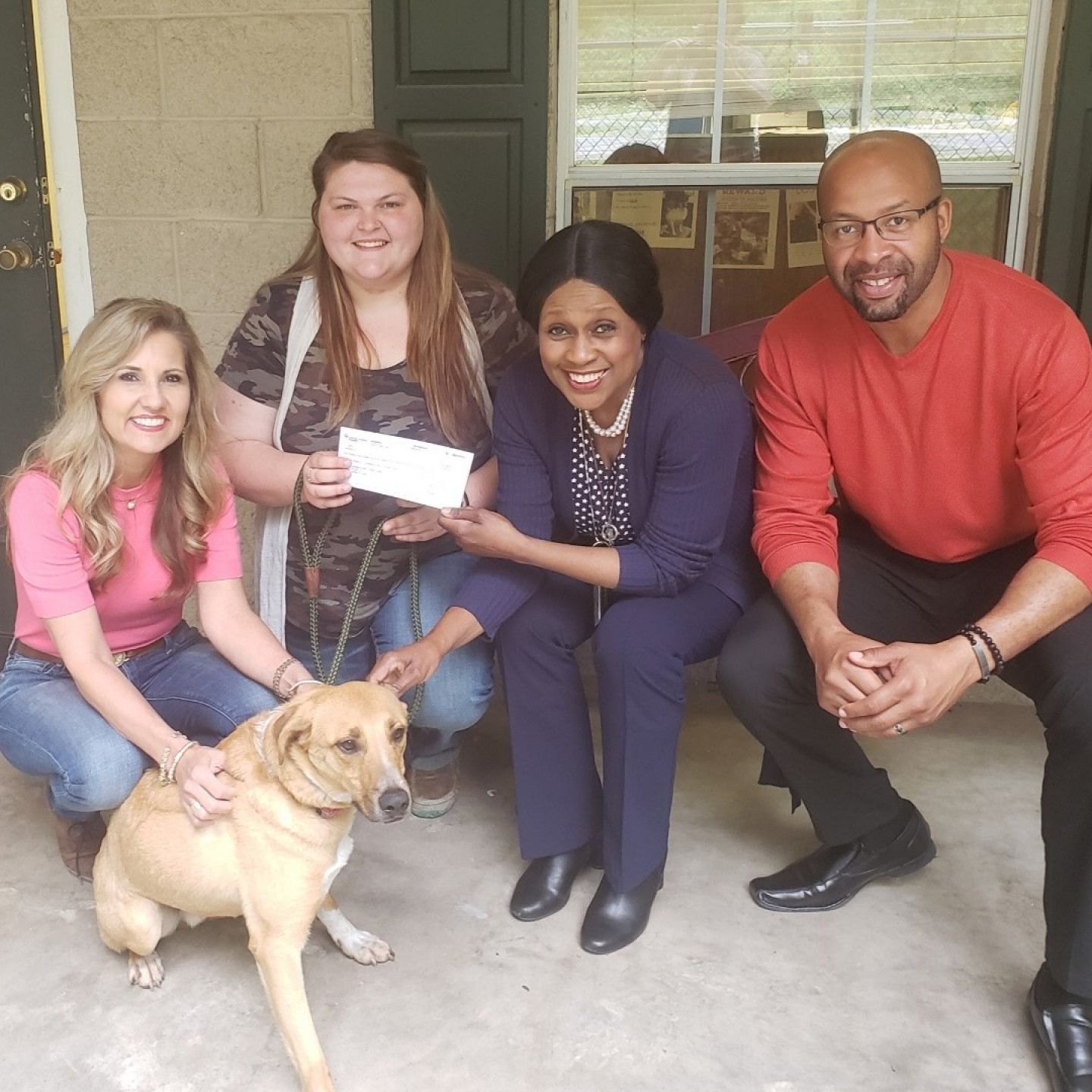 Four people and a dog pose on a porch. Woman holds a check, others smile.