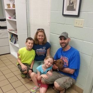 Family adopting a dog: two children, two adults, a brown dog in a room with a window and a bookshelf.
