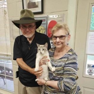 Couple holding a light-colored cat with blue eyes, posing indoors. Man wears a hat.