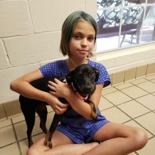 Girl with blue-tinted hair sits, holding a small black and tan puppy in a tiled room.