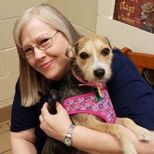 Woman smiling, holding a small dog with a pink harness. They are indoors.
