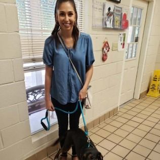 Woman in blue top, black pants, smiles while holding a dog on a leash in a building.