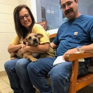 Couple seated, holding puppy, smiling. They sit indoors near a window.