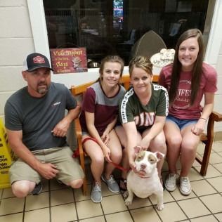 A family and dog pose on a bench inside. The dog is white with a red collar.