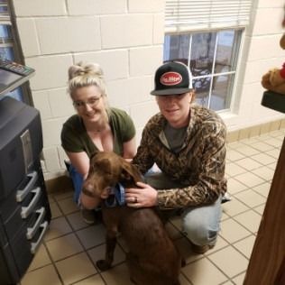 Two people kneel with a brown dog inside a building. The man pets the dog while the woman smiles.
