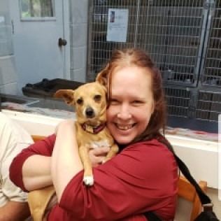 Woman in red shirt smiling, holding a small brown dog in an animal shelter.