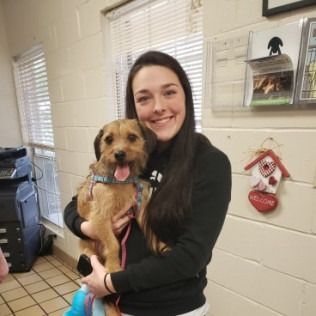 Woman smiles, holding a tan dog with its tongue out, inside a building.