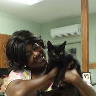 Woman smiling, holding a black cat indoors; a white air conditioner is in the background.