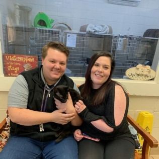 Two people holding a black cat in front of animal cages. They are smiling.