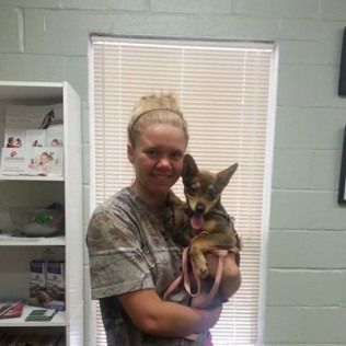 Woman holding a small brown dog, both smiling in front of a window. Inside, bookshelves are visible.