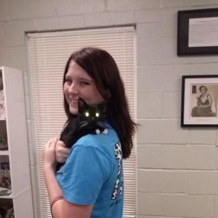 Woman with dark hair, wearing a blue shirt, holding a black cat on her shoulder; brick wall background.
