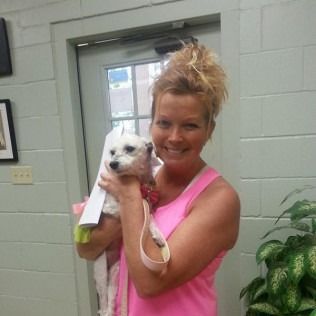 Woman in pink tank top holding a small white dog wrapped in a towel. Smiling indoors.