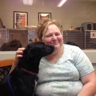 Woman smiling, petting a black dog. They are inside a pet shelter.