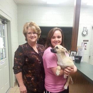 Two women with a puppy in an animal shelter; woman in pink shirt holding the dog.