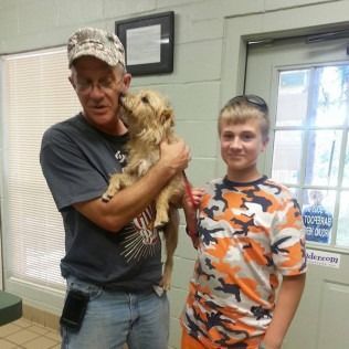 Man holding a small tan dog, kissing it. A boy stands next to them. Setting is inside with a door and window.