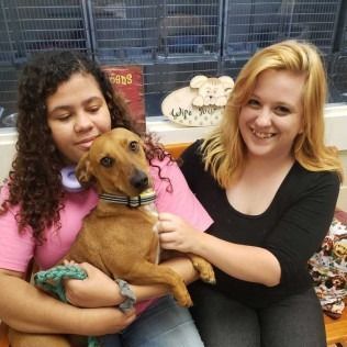 Two women smile, holding a brown dog in a shelter.