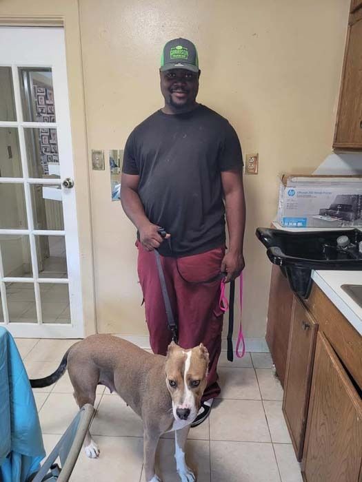 Man in black shirt and maroon pants stands with dog on leash in kitchen.