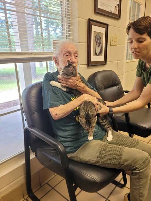 Man sitting with cat in his lap, another person assisting; indoors.