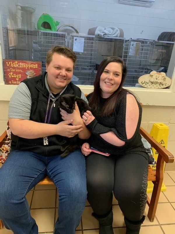 Two people smiling, holding a black cat in an animal shelter.