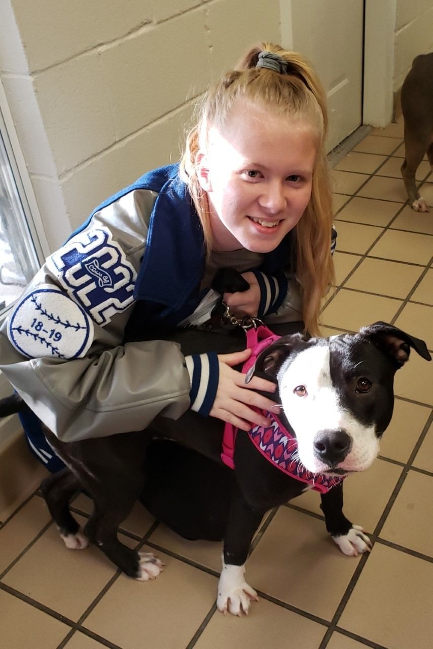 Woman in blue jacket smiles with a black and white dog wearing a pink harness and bandana in a tiled room.