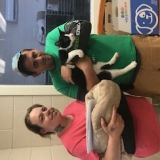 Two people holding a cat and a blanket in a brightly lit room. The people are smiling.