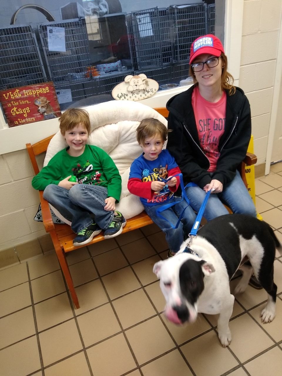 Family sits with a black and white dog on a bench. One child wears green, the other wears blue. The woman wears a red hat.