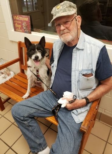 Man and dog sit on a bench. The dog is black and white, and the man wears a vest and hat.