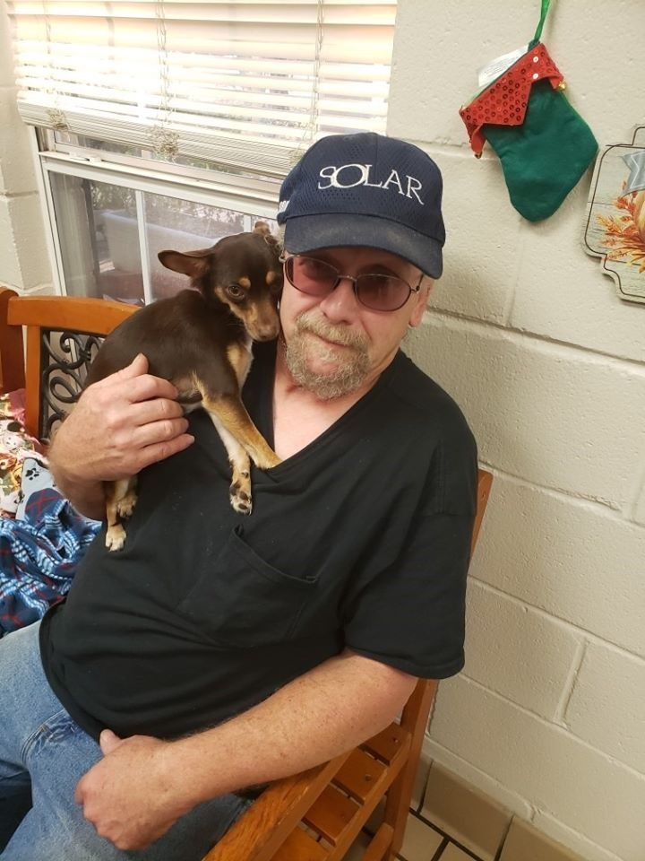Man in a hat holding a small brown dog, both looking at the camera.