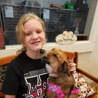 Girl with dog in shelter, dog licking girl's face. Both are smiling, with cages in the background.