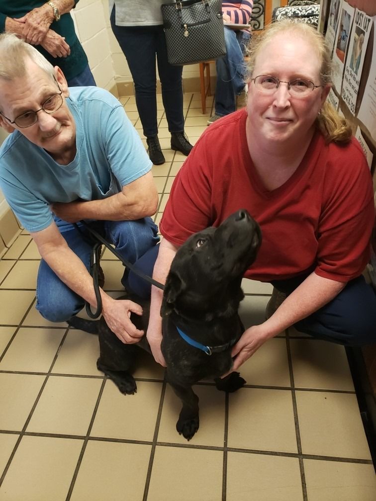 Man and woman kneeling with a black dog on a leash in a tile-floored room.