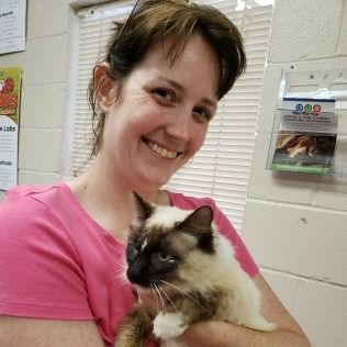 Woman holding a cat indoors, both smiling. The cat has a cream and brown coat; the woman wears pink.