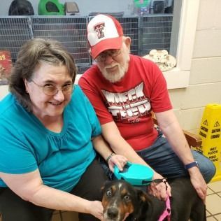 A woman and man smiling, petting a dog on a leash inside a building.