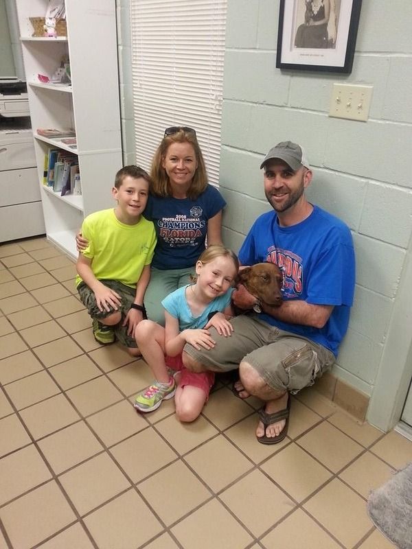 Family with children, holding a small dog, smiling. They're indoors near shelves.